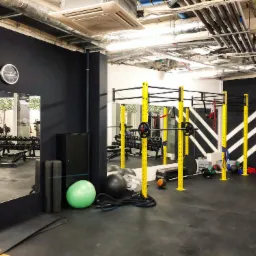 Wnętrze siłowni z czarnymi ścianami, żółtymi elementami konstrukcyjnymi, widoczne lustra, sprzęt do ćwiczeń, piłki, liny i sztangi.