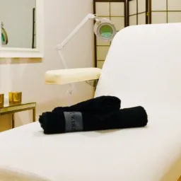 Biały stół do zabiegów z czarnymi ręcznikami, lampą powiększającą i złotymi świecznikami w tle.