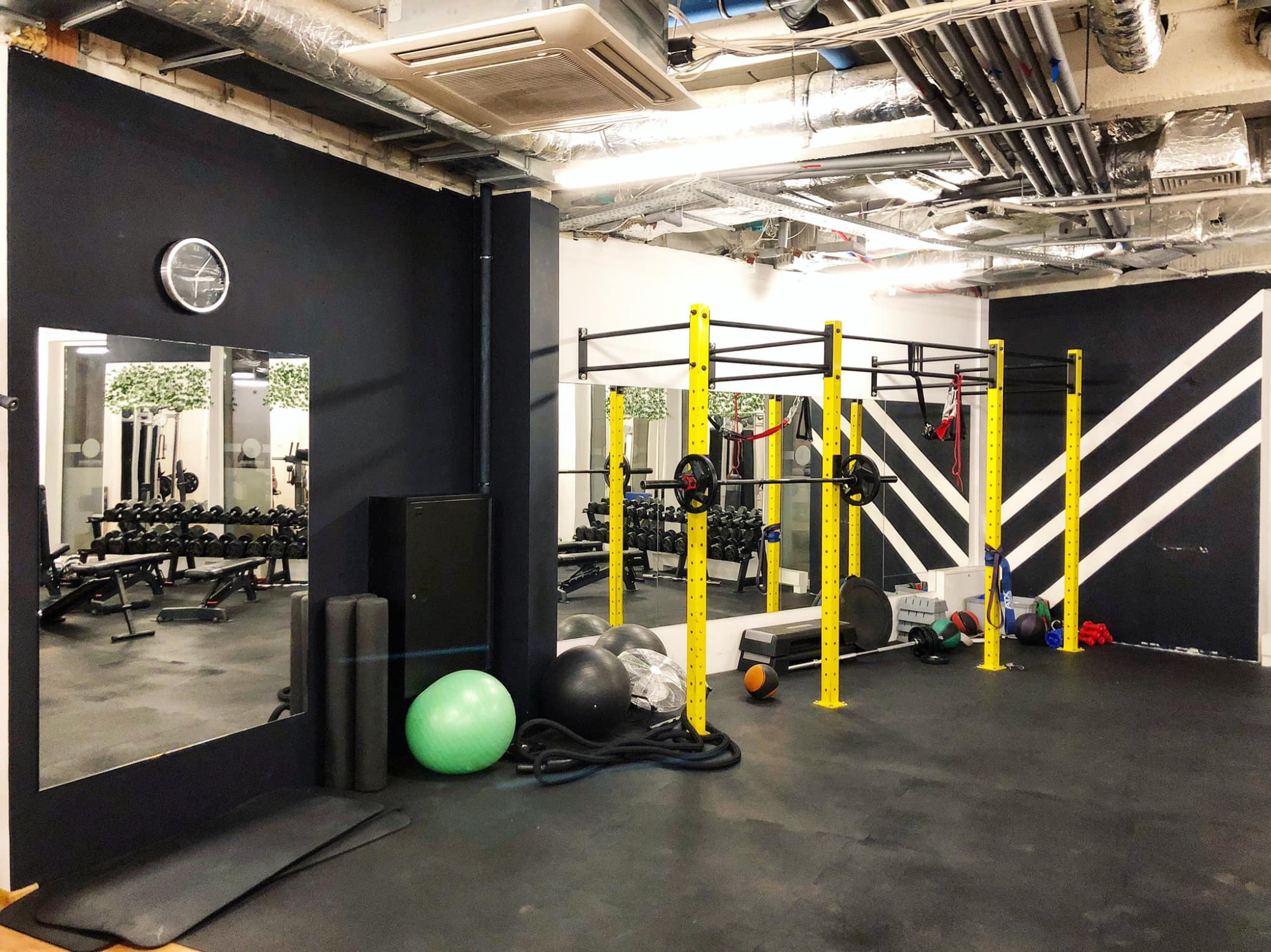 Wnętrze siłowni z czarnymi ścianami, żółtymi elementami konstrukcyjnymi, widoczne lustra, sprzęt do ćwiczeń, piłki, liny i sztangi.