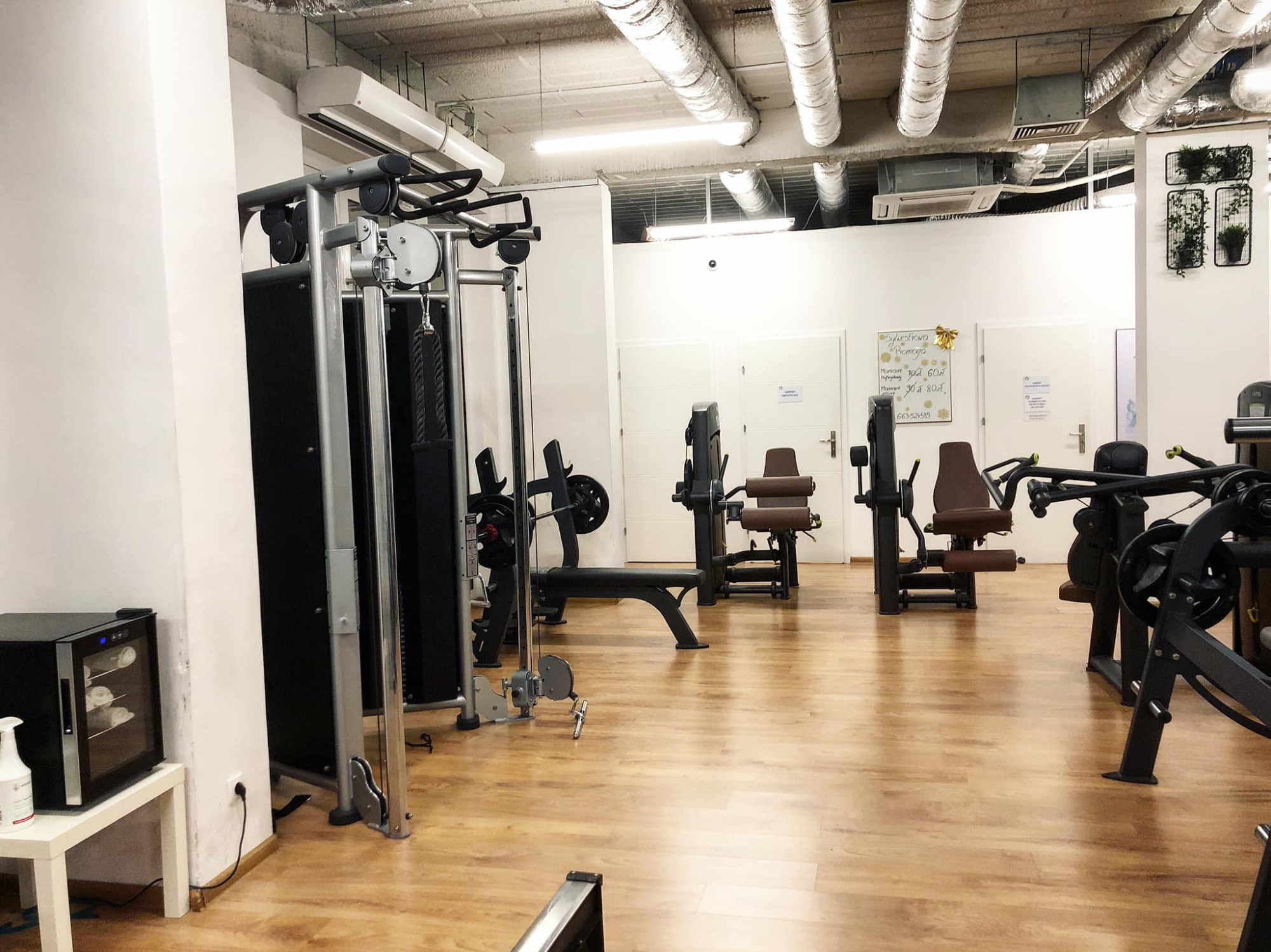 Wnętrze przestronnej siłowni z różnorodnym sprzętem do ćwiczeń, w tle białe ściany z dekoracjami roślinnymi i tablicą informacyjną.