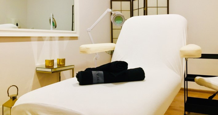 Biały stół do zabiegów z czarnymi ręcznikami, lampą powiększającą i złotymi świecznikami w tle.