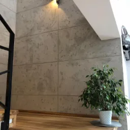 Ściana wykończona betonem architektonicznym w odcieniach szarości, oświetlona kinkietem, z drewnianymi schodami z podświetleniem LED i rośliną doniczkową w białej osłonce.