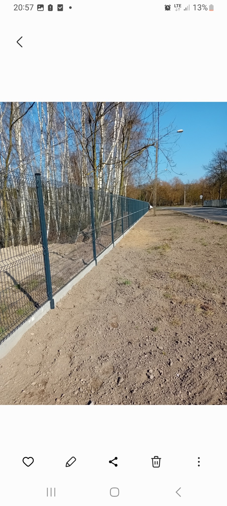 Długi, ciemnoszary system ogrodzeniowy z siatki panelowej na betonowej podmurówce, biegnący wzdłuż piaszczystego terenu i drzew z nagimi gałęziami w tle.