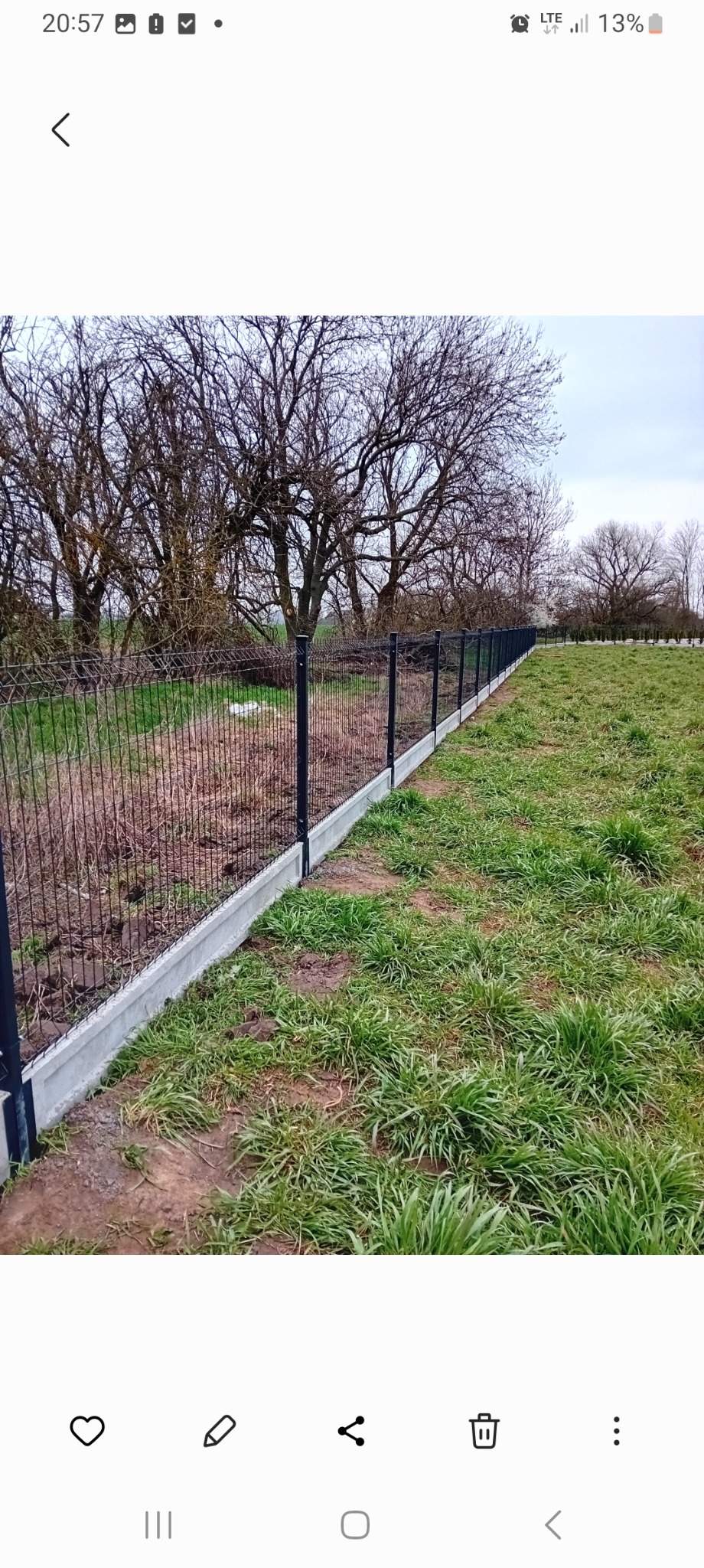 Długi odcinek czarnego ogrodzenia z siatki na betonowej podmurówce, oddzielający teren z suchą trawą i drzewami bez liści w tle.