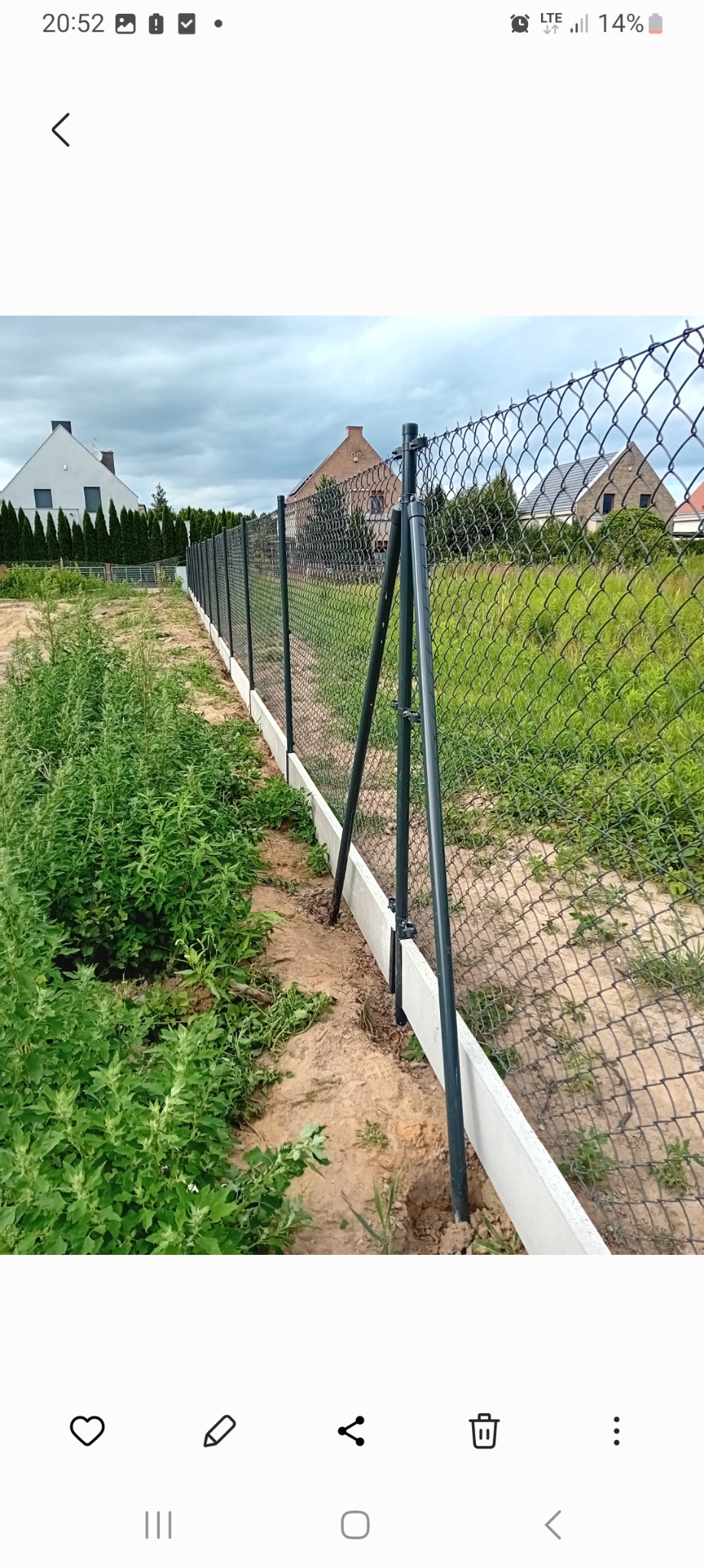 Długa linia ciemnej siatki ogrodzeniowej na betonowej podmurówce, wsparta na metalowych słupkach z podporami, oddzielająca działki porośnięte trawą i chwastami, z domami w tle.