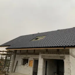 Dom murowany w trakcie budowy z czarną dachówką, widoczne rusztowanie i tablica informacyjna na ścianie z białych bloczków.