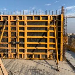 Żółte szalunki z widocznymi elementami mocującymi i zbrojeniem na budowie, w tle budynki i dźwig.