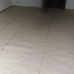 Przygotowane podłoże pod nową podłogę, zabezpieczone arkuszami ochronnymi przed dalszymi pracami remontowymi, widoczne ściany w stanie surowym oraz otwarte drzwi do kolejnego pomieszczenia.