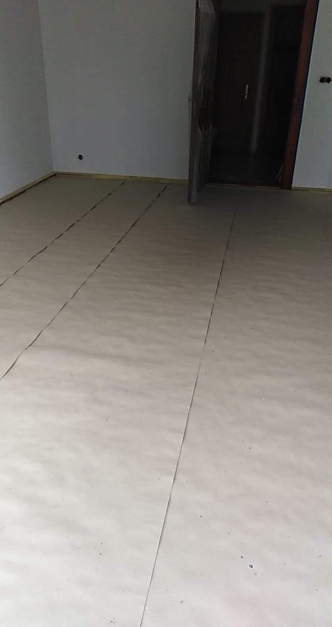 Przygotowane podłoże pod nową podłogę, zabezpieczone arkuszami ochronnymi przed dalszymi pracami remontowymi, widoczne ściany w stanie surowym oraz otwarte drzwi do kolejnego pomieszczenia.