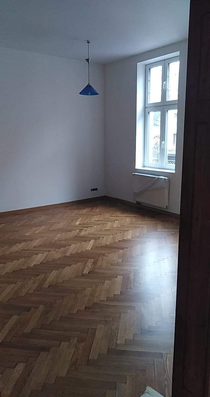 Pokój po renowacji: parkiet ułożony w jodełkę, białe ściany, okno z grzejnikiem, niebieska lampa wisząca.