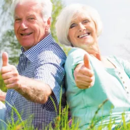 Uśmiechnięta para seniorów siedzi na trawie, opierając się plecami o siebie i pokazując kciuki w górę w słoneczny dzień.