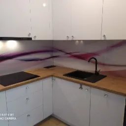 Nowoczesna kuchnia z białymi frontami szafek, drewnianym blatem i czarnym zlewem oraz płytą indukcyjną. Ściana nad blatem zdobiona grafiką w odcieniach fioletu i bieli.