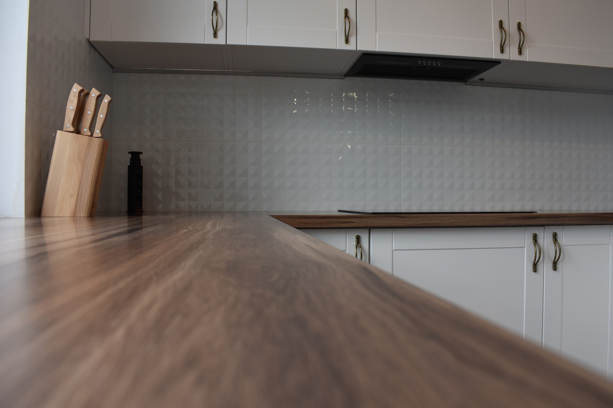 Nowoczesna kuchnia z białymi szafkami i drewnianym blatem, widok z bliska na fakturę blatu, w tle stojak na noże i białe płytki ścienne z subtelnym wzorem.