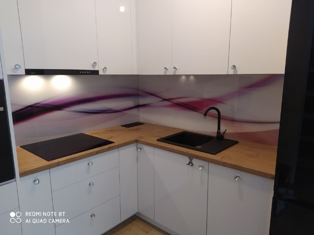 Nowoczesna kuchnia z białymi frontami szafek, drewnianym blatem i czarnym zlewem oraz płytą indukcyjną. Ściana nad blatem zdobiona grafiką w odcieniach fioletu i bieli.