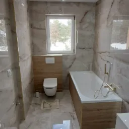 Wykończona łazienka z marmurowymi płytkami, drewnopodobną obudową wanny i podwieszaną toaletą z drewnianym panelem, okno z widokiem na zimowy krajobraz.