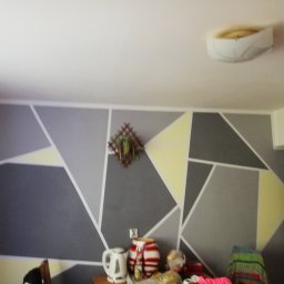 Twardowski mateusz - Ściana z geometrycznym wzorem w odcieniach szarości i żółci, ozdobiona dekoracją roślinną. Na pierwszym planie widoczne przedmioty na stole.