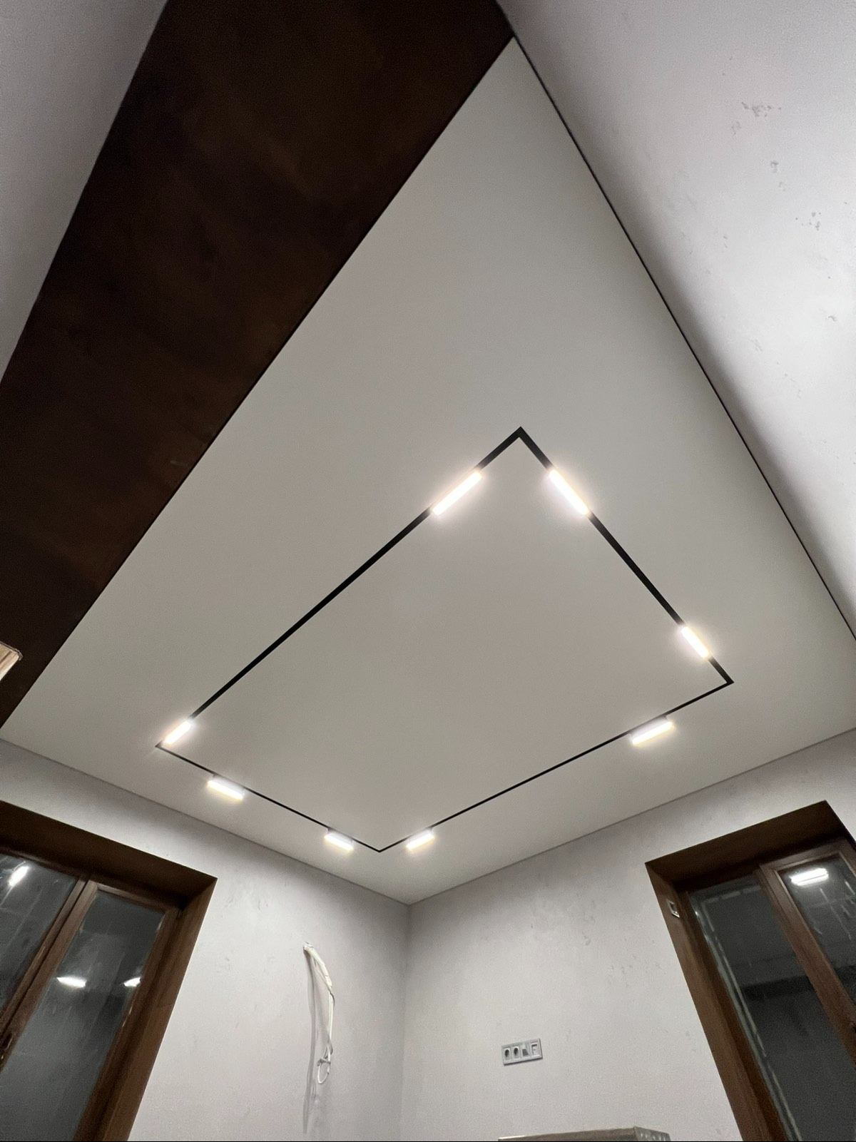 Biały sufit napinany z czarną ramą i wbudowanym oświetleniem LED, widok z dołu, okna w tle. Nowoczesny design wnętrza.