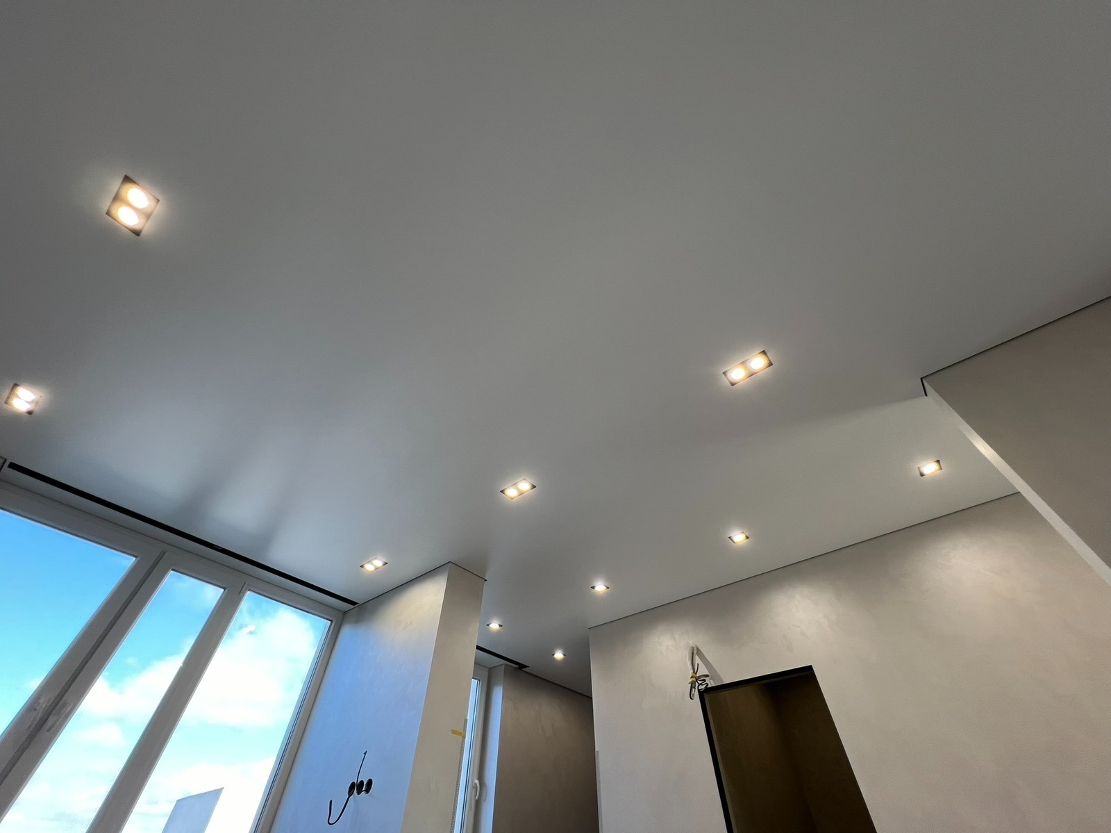 Biały sufit napinany z równomiernie rozmieszczonymi, kwadratowymi lampami LED w nowoczesnym wnętrzu. Widok z dołu, jasne światło dzienne.