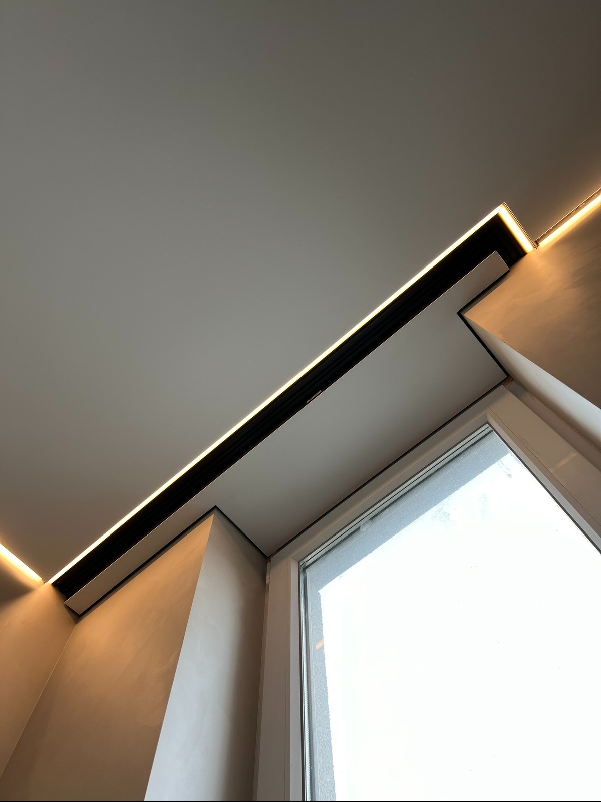 Nowoczesny sufit podwieszany z oświetleniem LED wzdłuż okna. Minimalistyczny design, ciepłe światło. Kątowe ujęcie podkreśla geometrię wnętrza.