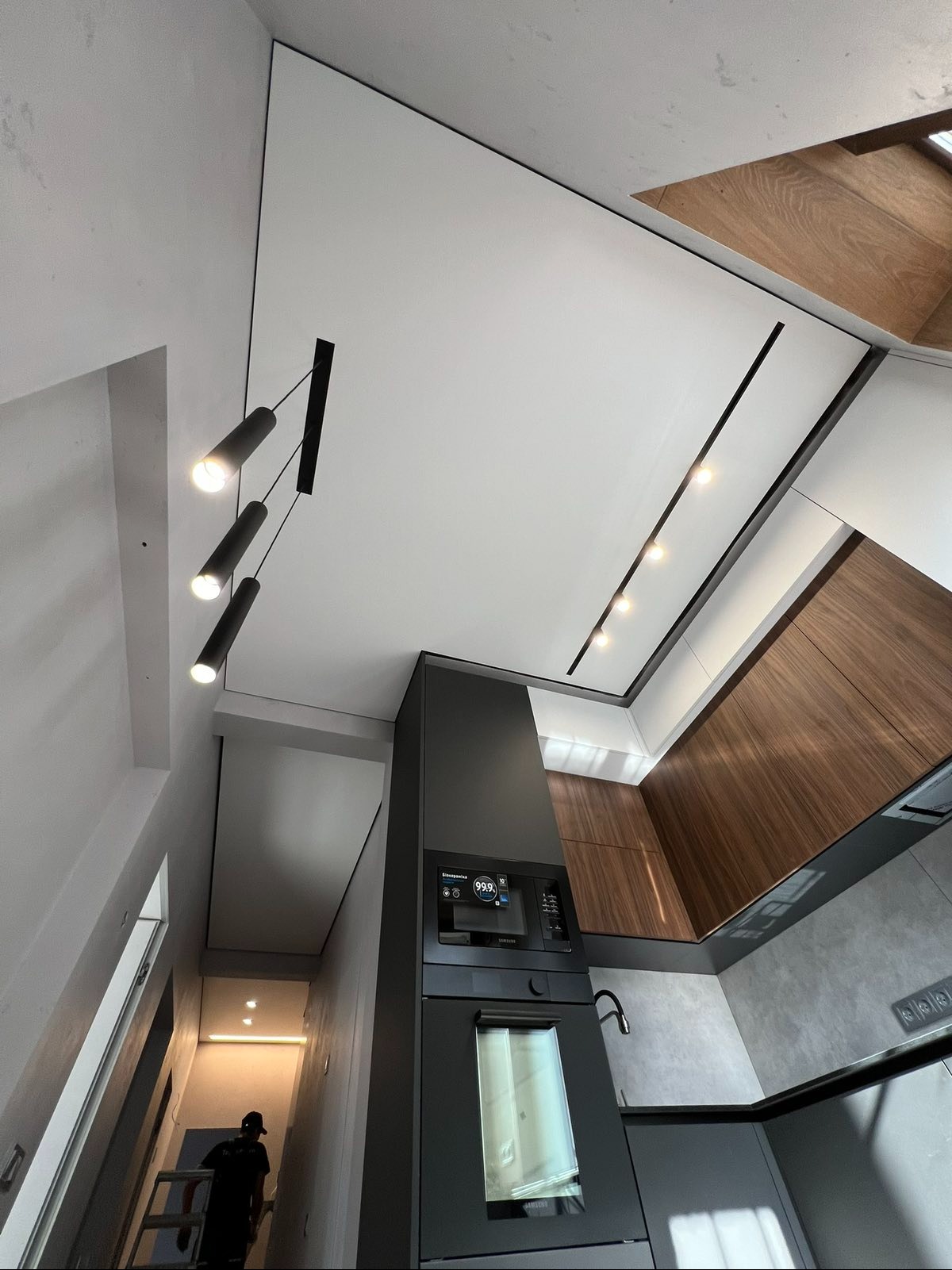Nowoczesny sufit napinany z oświetleniem punktowym w kuchni, widok z dołu. Czarny panel z lampami. Elementy drewniane i szare. Osoba na drabinie w tle.