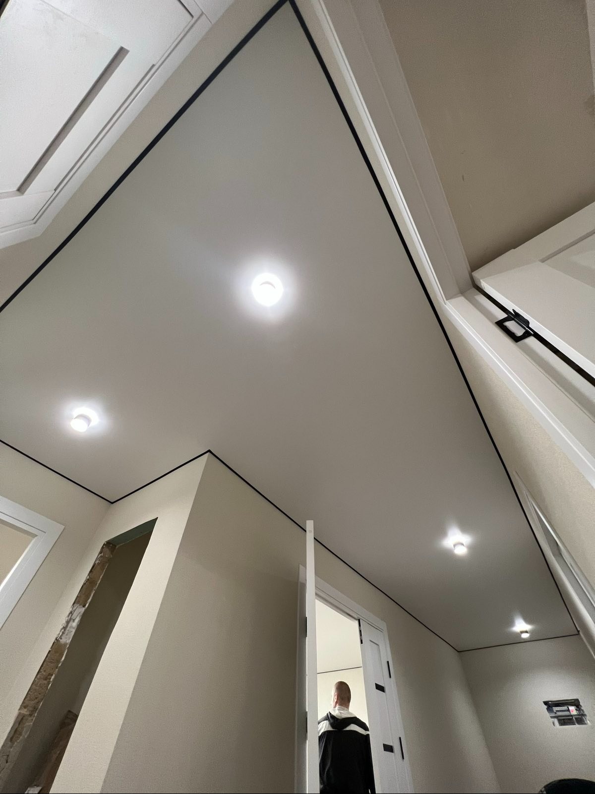 Biały sufit napinany z czarnym obramowaniem i wbudowanymi lampami, widok z dołu w trakcie wykańczania wnętrza, z otwartymi drzwiami i widoczną osobą.