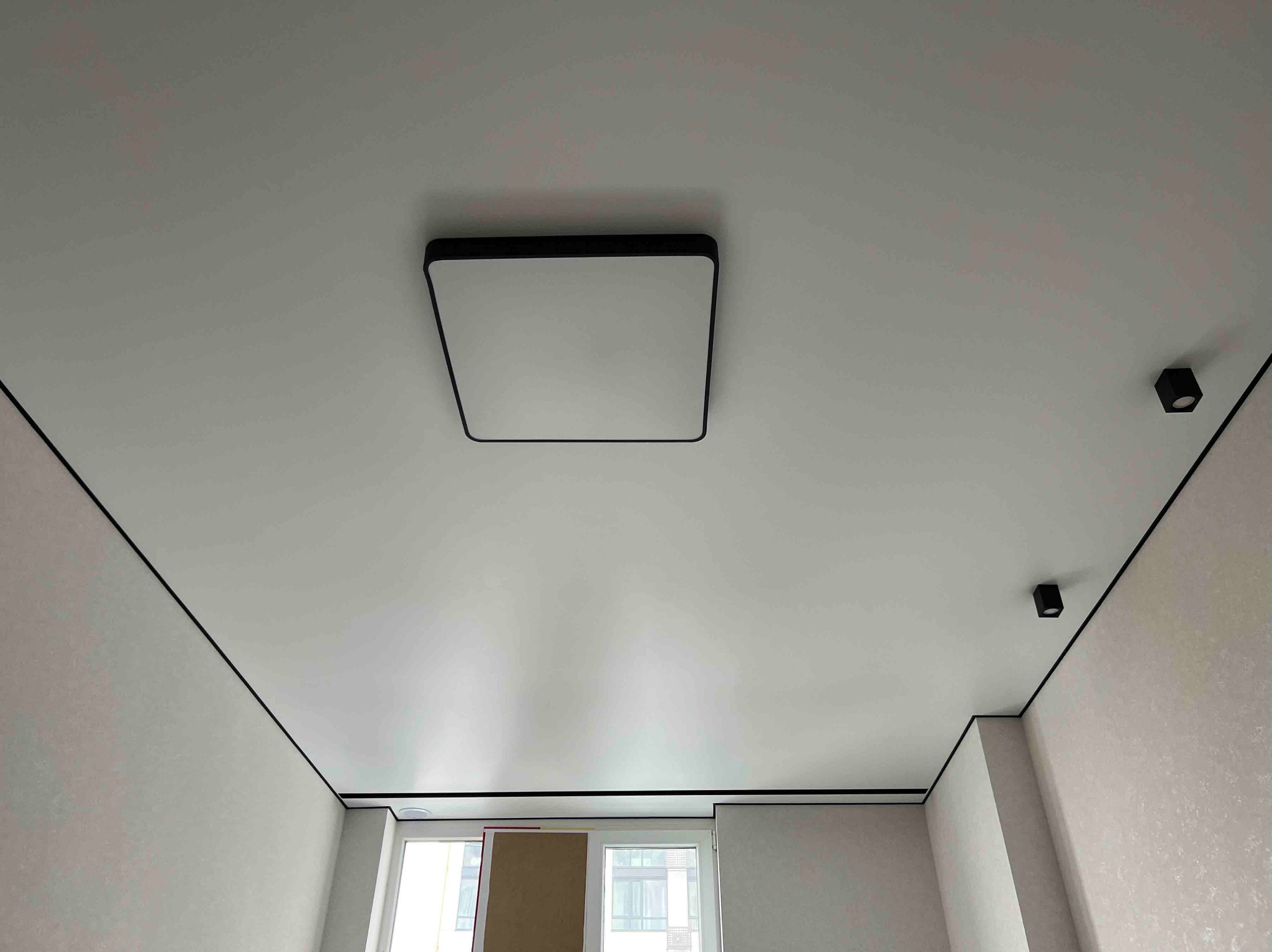 Biały sufit napinany z czarnymi listwami i nowoczesnym oświetleniem LED. Kwadratowa lampa centralna i małe reflektory punktowe. Widok z dołu, wykończone wnętrze.
