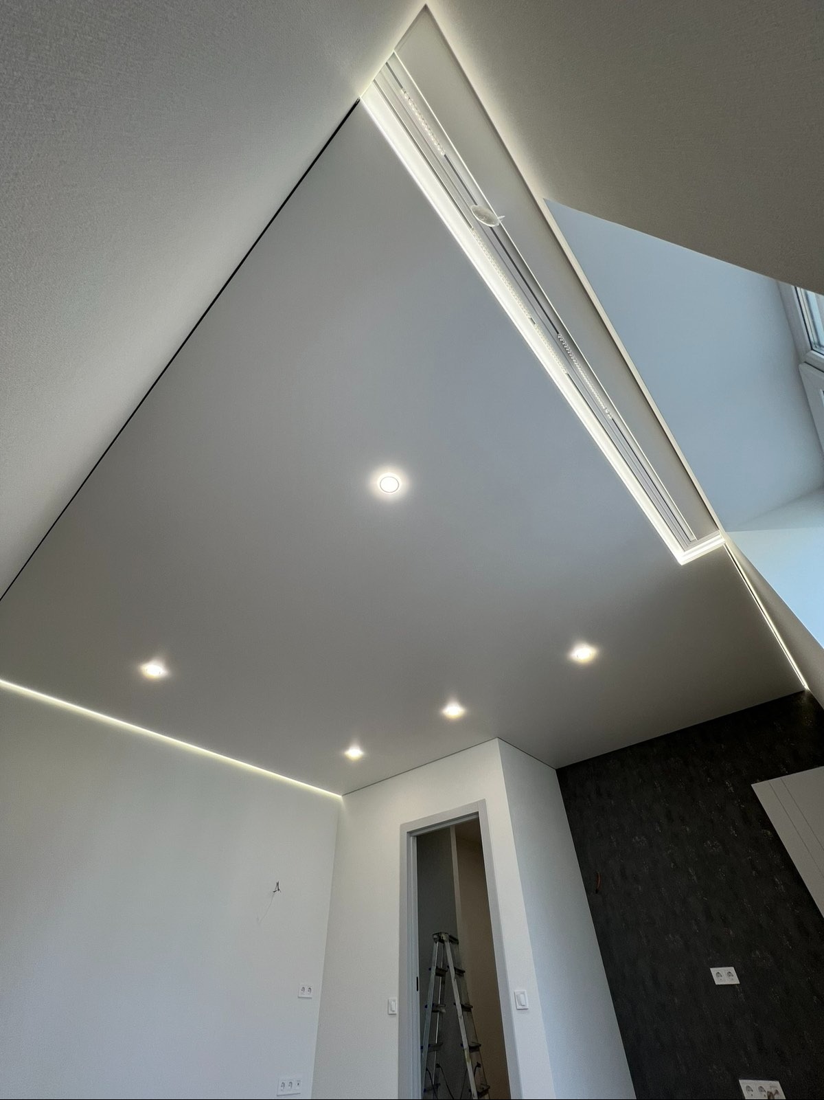 Nowoczesny sufit napinany z oświetleniem LED i punktowymi lampami w wykończonym wnętrzu. Widoczna drabina. Białe ściany i fragment ściany z ciemną tapetą.