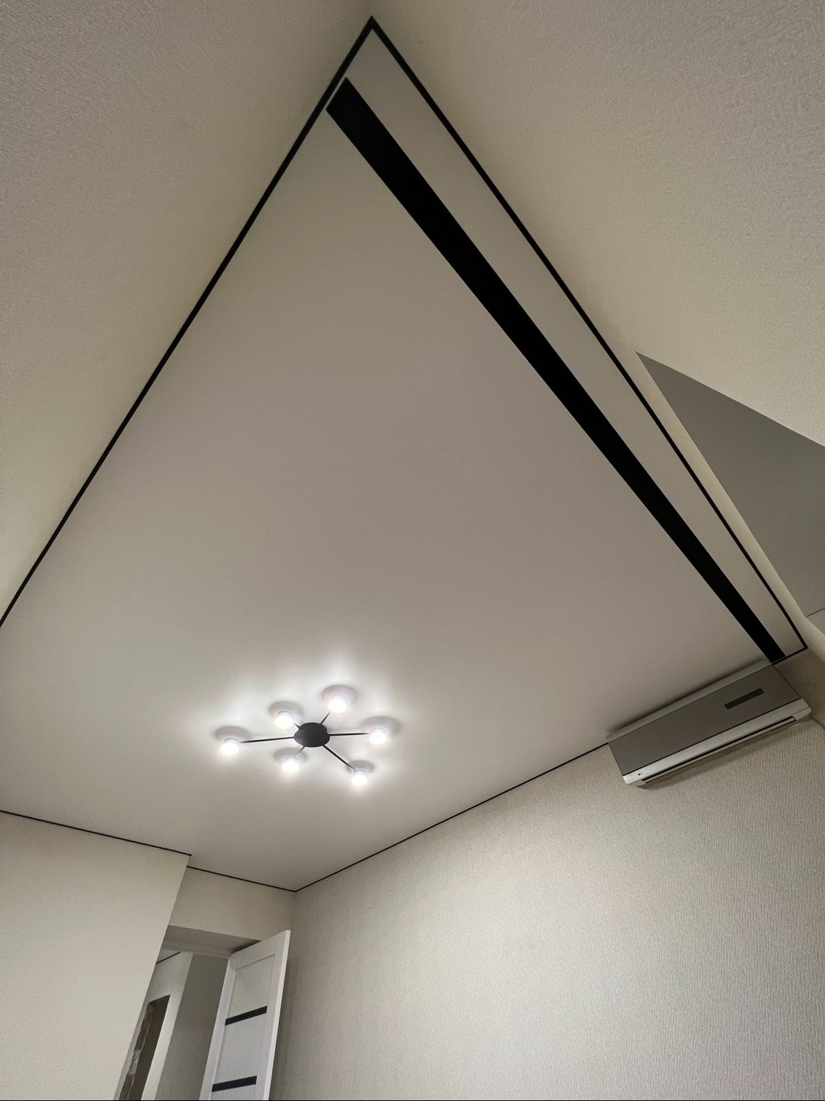 Biały sufit napinany z czarnymi listwami, oświetleniem punktowym i klimatyzacją. Ujęcie z dołu, widoczne drzwi w tle.