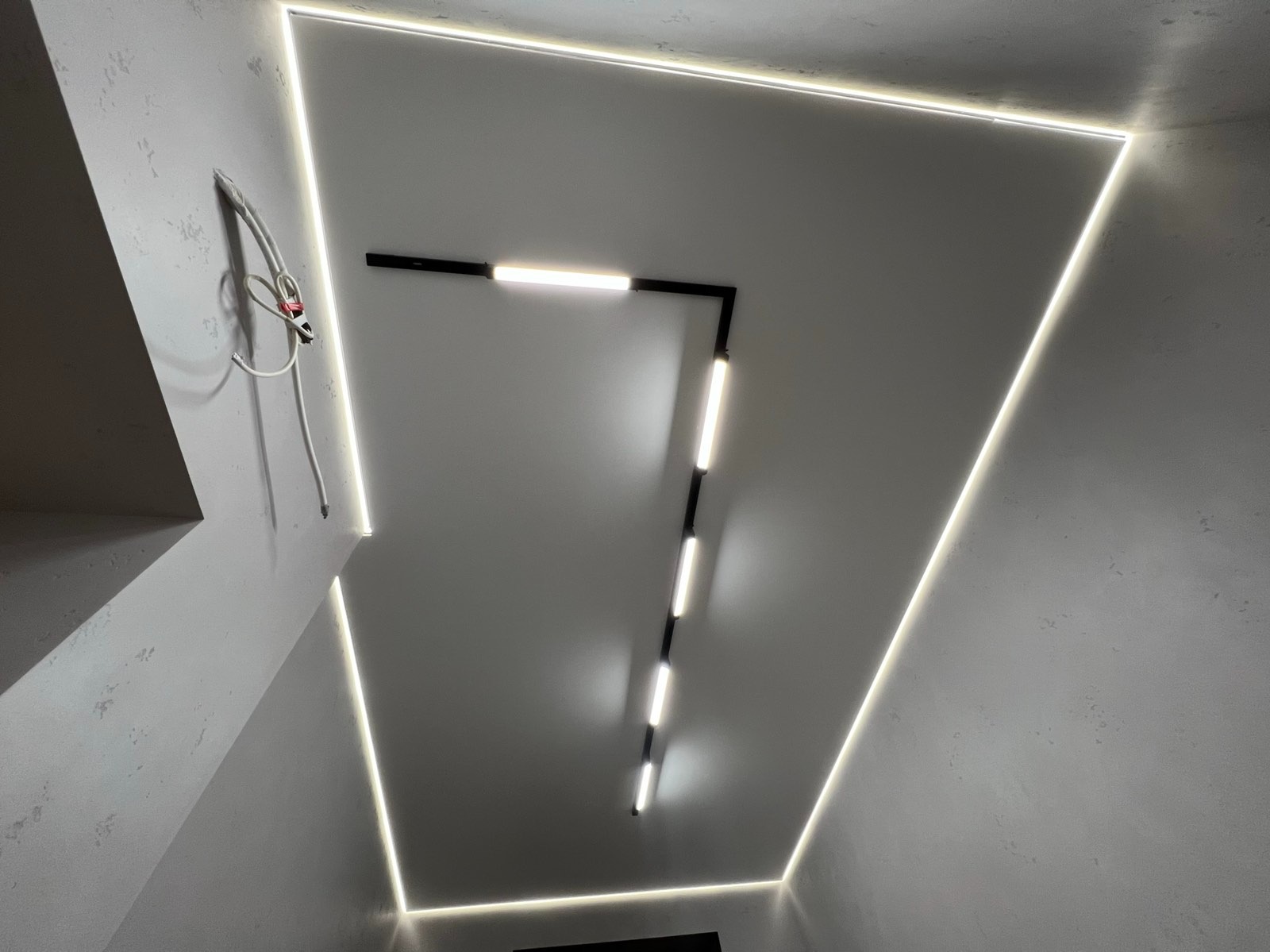 Nowoczesny sufit z liniowym oświetleniem LED w kształcie trapezu i prostokątną lampą, minimalistyczny design, szare ściany z fakturą, kabel wystający ze ściany.