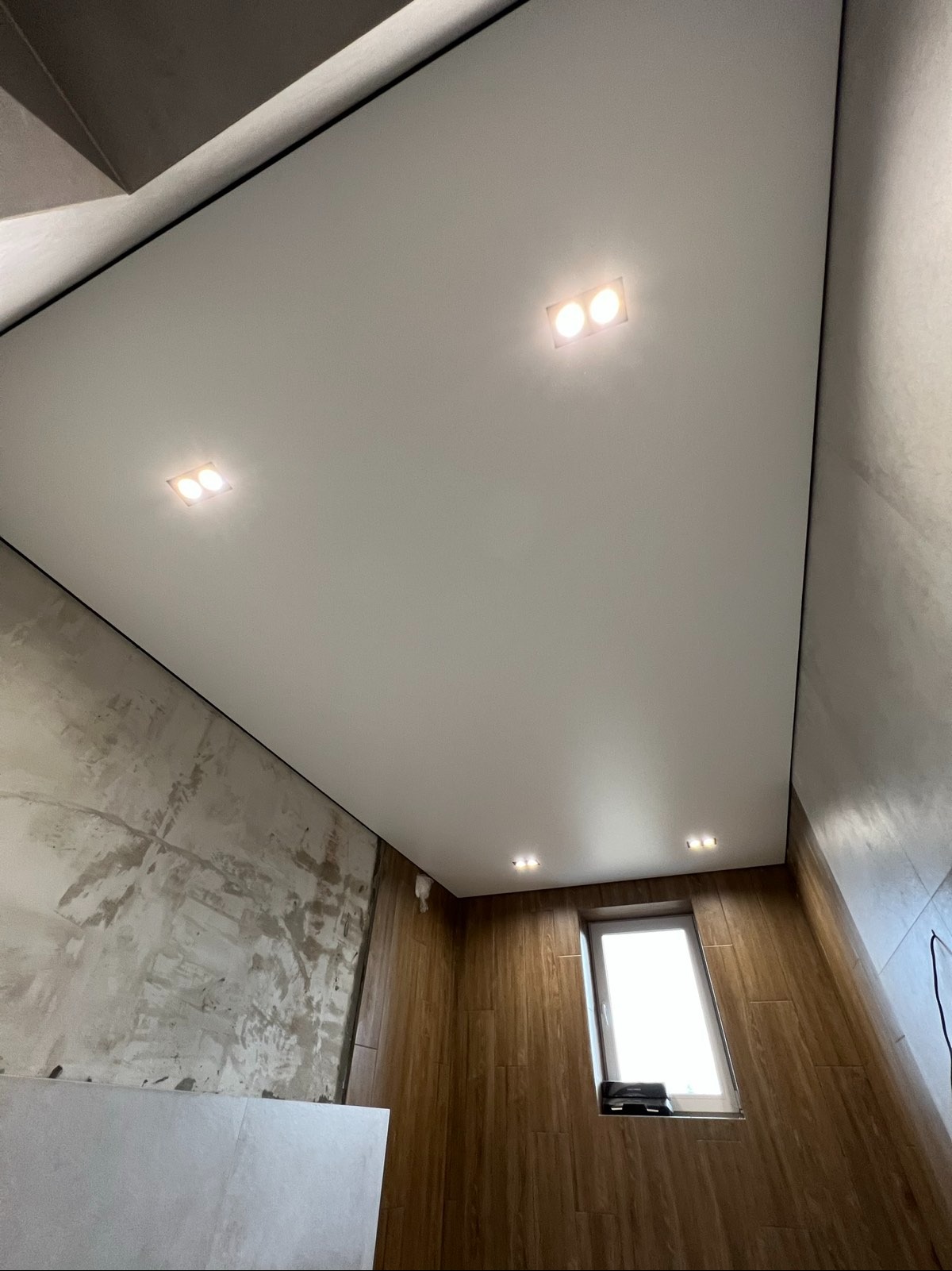 Biały sufit napinany z czterema punktami świetlnymi w łazience z wykończonymi drewnem ścianami i oknem, widok z dołu.