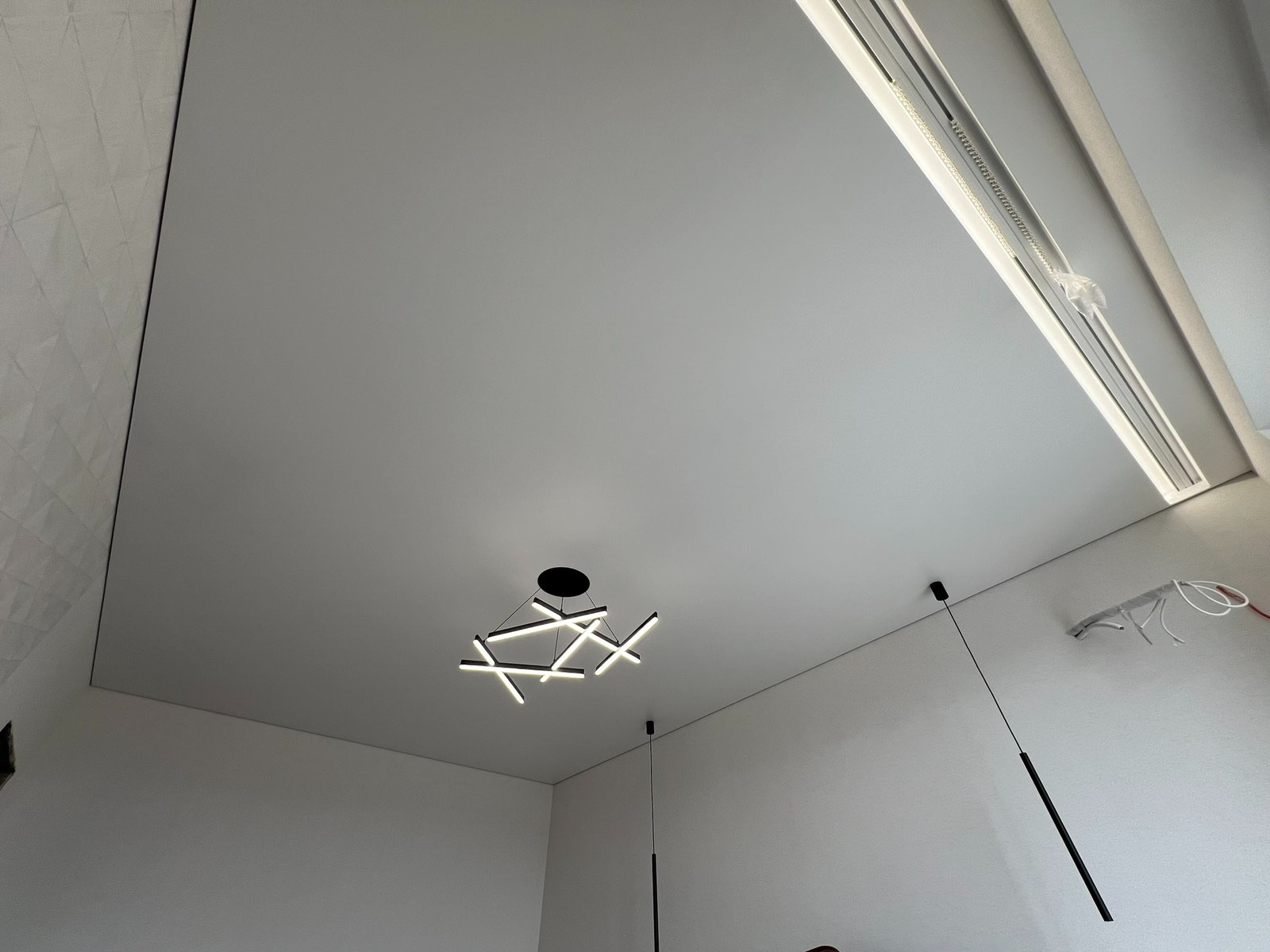 Biały sufit napinany z oświetleniem LED i nowoczesnymi lampami wiszącymi. Widoczny fragment ściany z dekoracyjną fakturą. Minimalistyczne wnętrze.