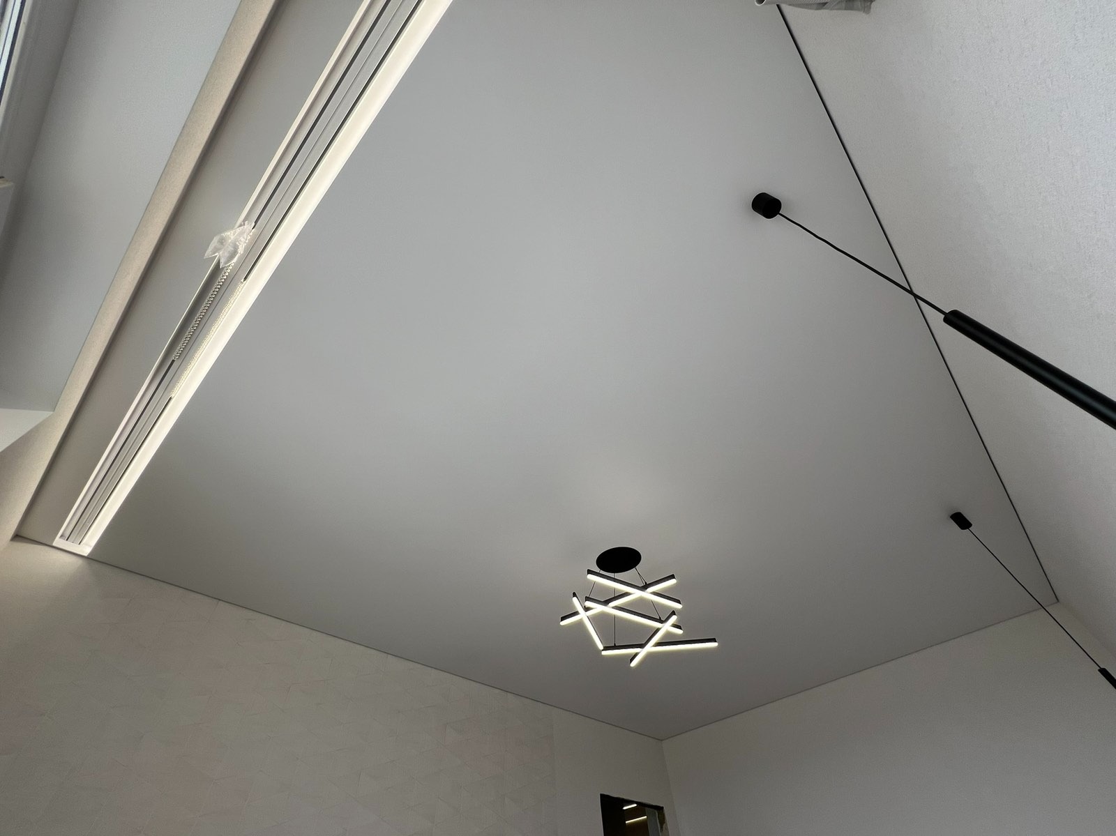 Biały sufit napinany z oświetleniem LED, nowoczesny design, widoczne mocowania lampy wiszącej. Minimalistyczne wykończenie wnętrza.