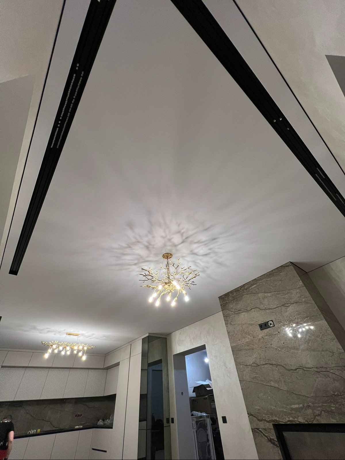 Nowoczesny sufit napinany z czarnymi listwami LED i designerskim żyrandolem w kształcie gałęzi. Jasne wnętrze z elementami dekoracyjnymi na ścianie.