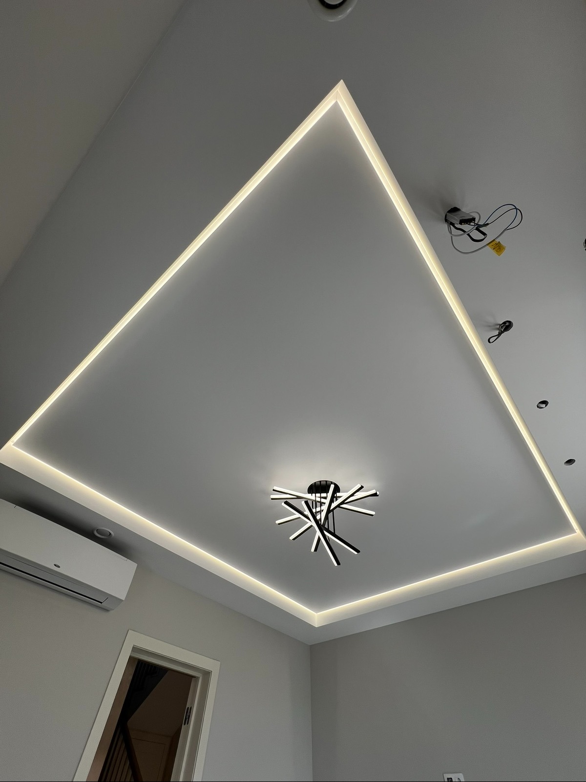 Nowoczesny sufit podwieszany z oświetleniem LED w kształcie kwadratu i designerską lampą. Widoczny klimatyzator i fragment drzwi. Minimalistyczny styl wykończenia wnętrza.