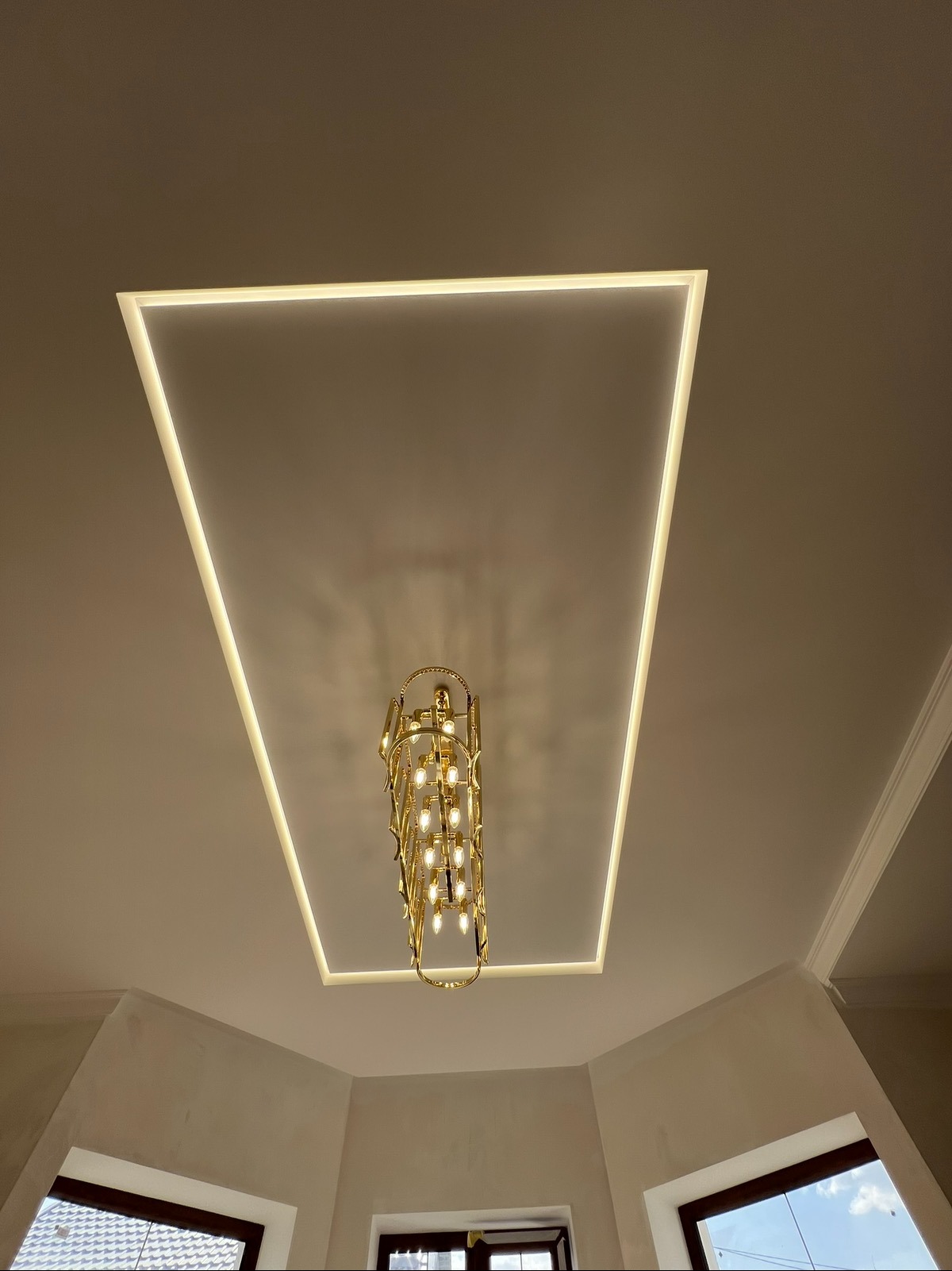 Beżowy sufit z kwadratowym oświetleniem LED i złotym, kaskadowym żyrandolem. Widok z dołu na okna i ściany w neutralnych barwach.