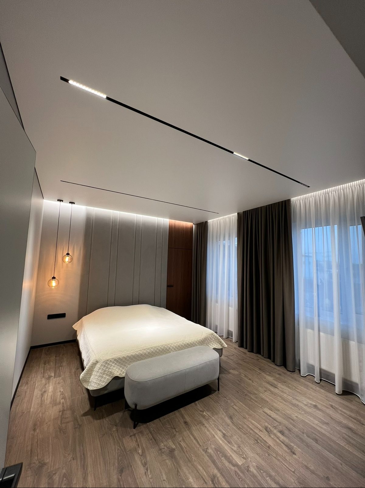 Nowoczesna sypialnia z sufitem napinanym i liniowym oświetleniem LED. Parkiet w odcieniach brązu i beżowe zasłony dopełniają minimalistyczny wystrój.