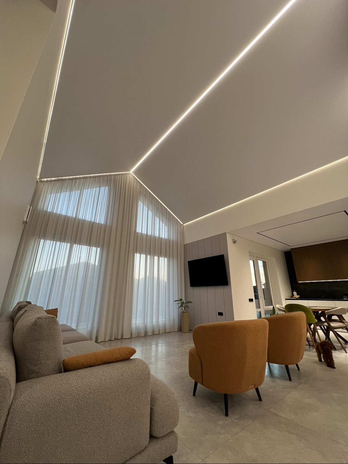 Nowoczesny salon z sufitem napinanym z podświetleniem LED, jasnymi zasłonami i minimalistycznym wystrojem. Widoczny fragment sofy i foteli.