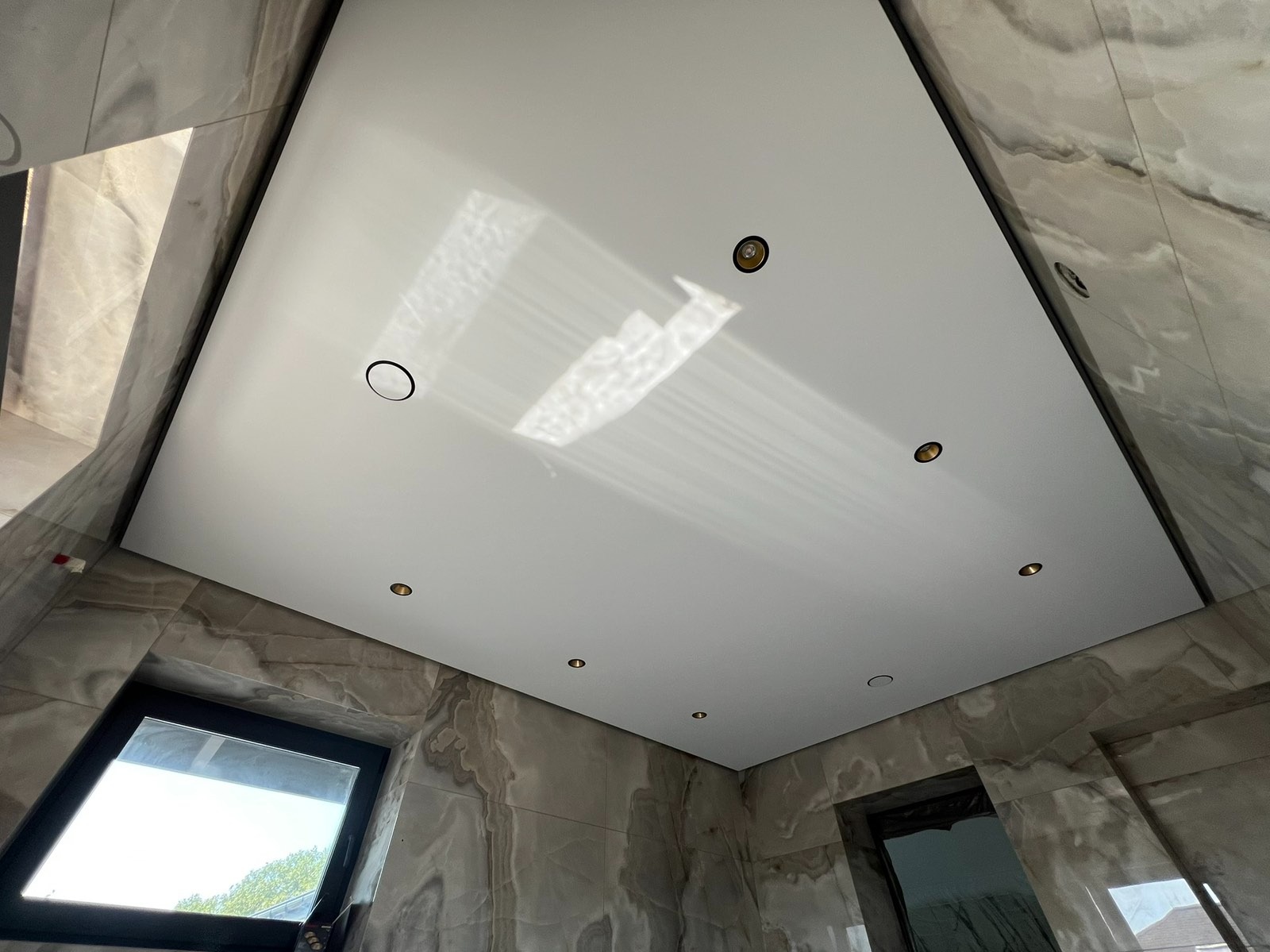 Biały sufit napinany z oświetleniem punktowym w łazience wykończonej marmurem, widok z dołu, z odbiciem światła w połyskliwej powierzchni.