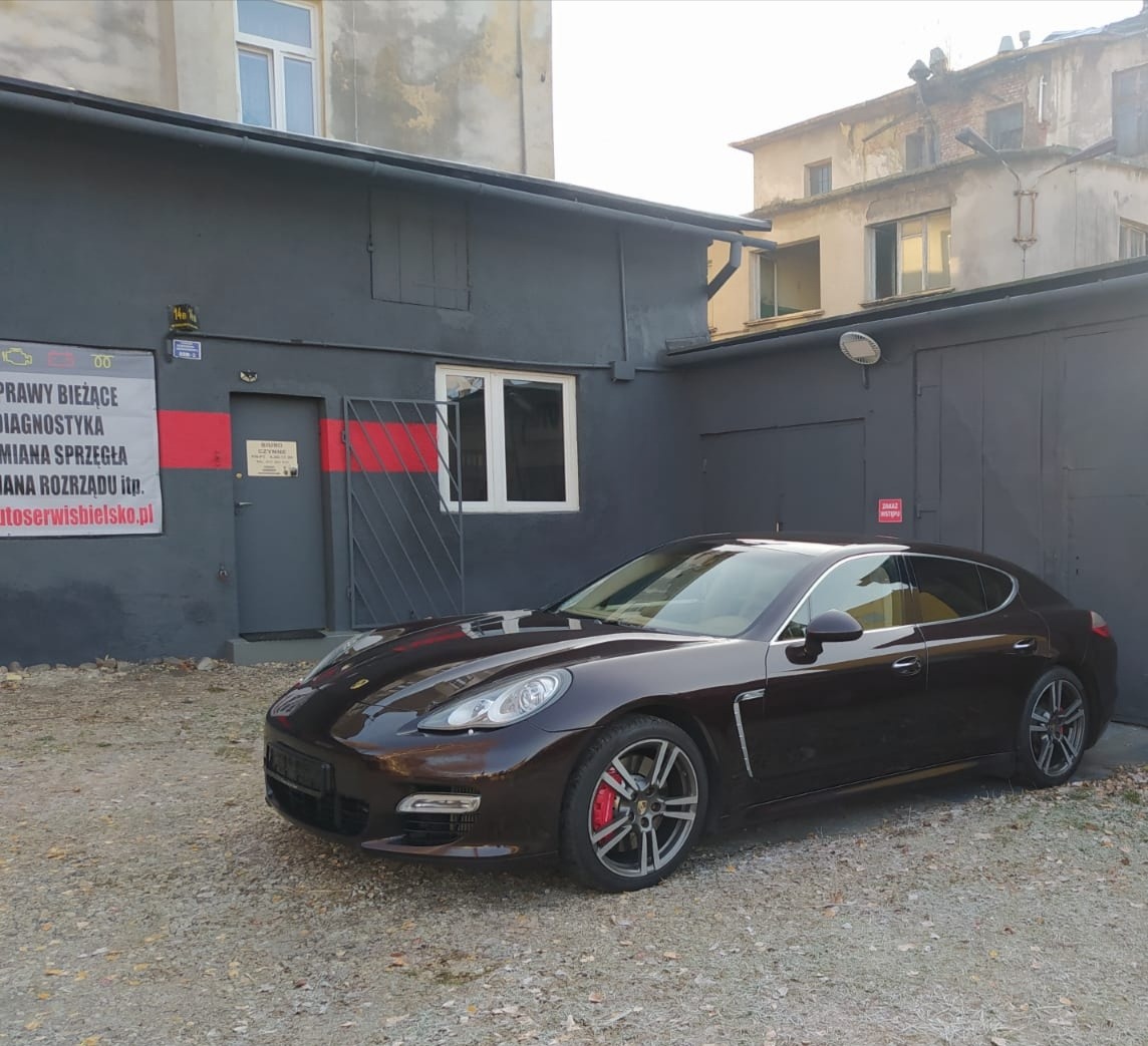 Ciemnobrązowa Porsche Panamera zaparkowana przed warsztatem samochodowym z szyldem informującym o naprawach bieżących, diagnostyce, wymianie sprzęgła i rozrządu, widoczny adres strony internetowej...