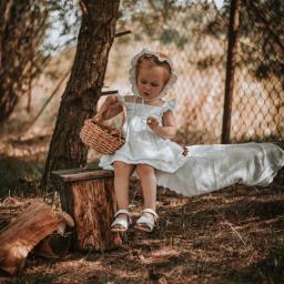 Ulyana Palamar - Mała dziewczynka w białej sukience i czepku siedzi na drewnianym pieńku w lesie, trzymając wiklinowy koszyk z zawartością i dotykając go palcem.
