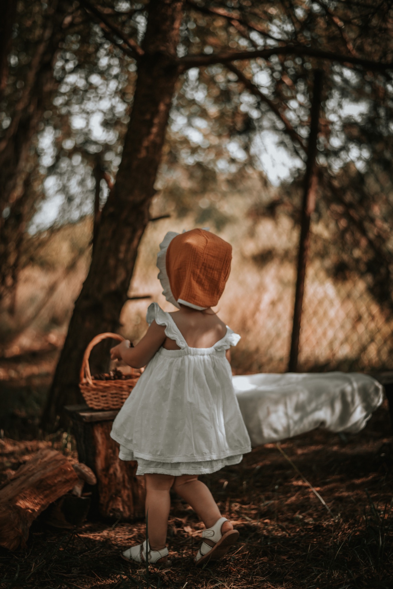Dziewczynka w białej sukience i pomarańczowym czepku, odwrócona tyłem, trzyma wiklinowy koszyk, spaceruje w lesie, w tle rozmyte drzewa i białe poszycie.
