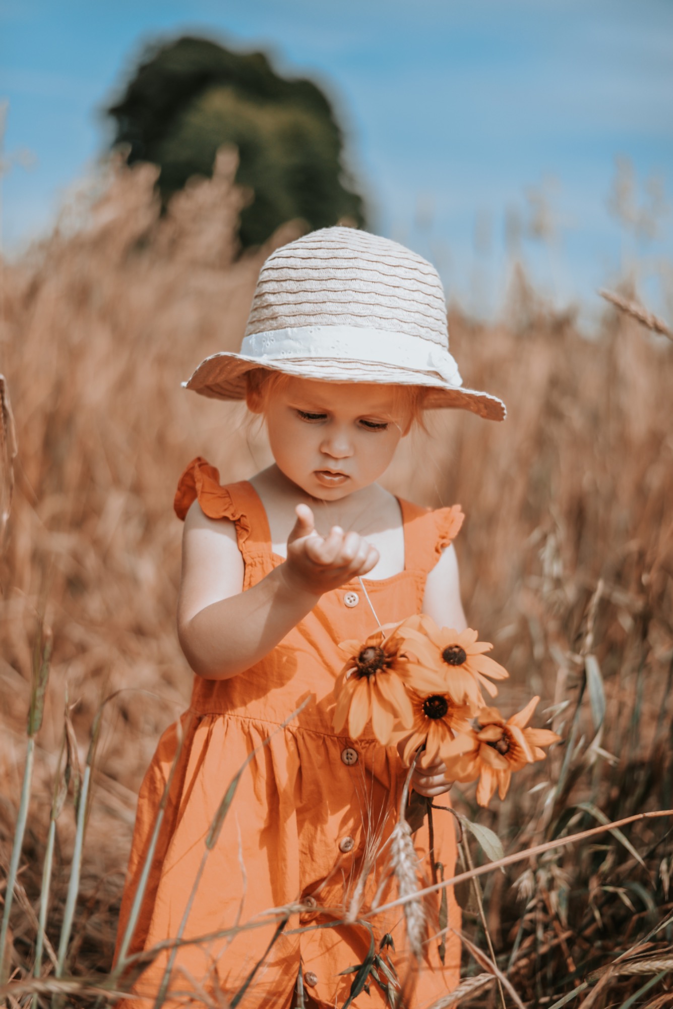 Dziewczynka w słomkowym kapeluszu i pomarańczowej sukience trzyma bukiet rudbekii w polu zboża, skupiona na czymś w dłoni.