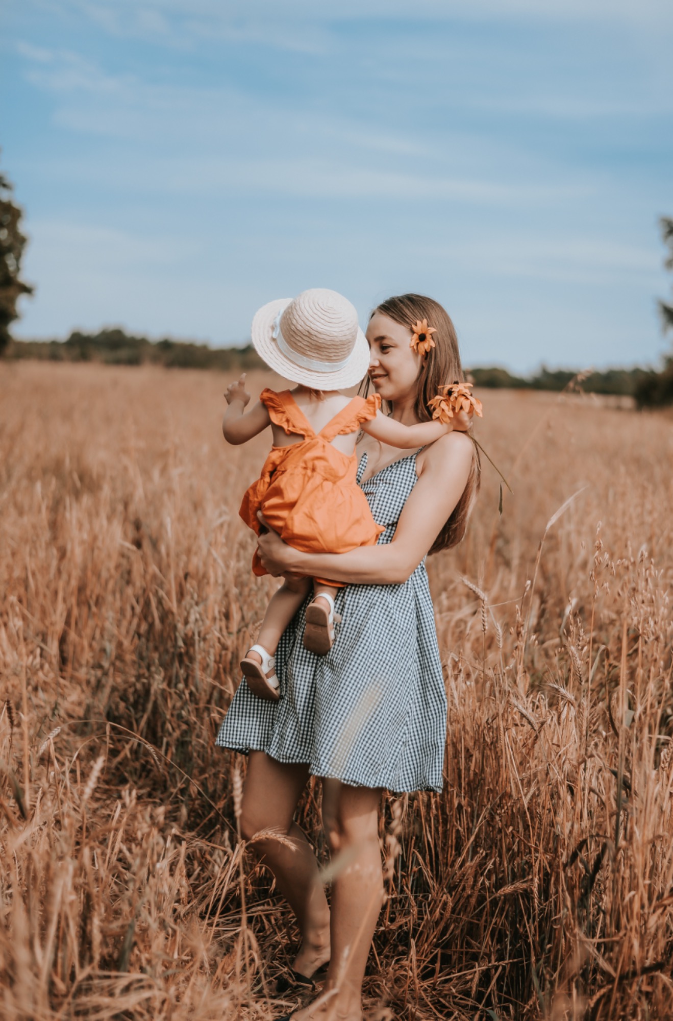 Matka trzymająca małą córeczkę w słomkowym kapeluszu na tle pola zboża, kobieta ma we włosach wpięty słonecznik.
