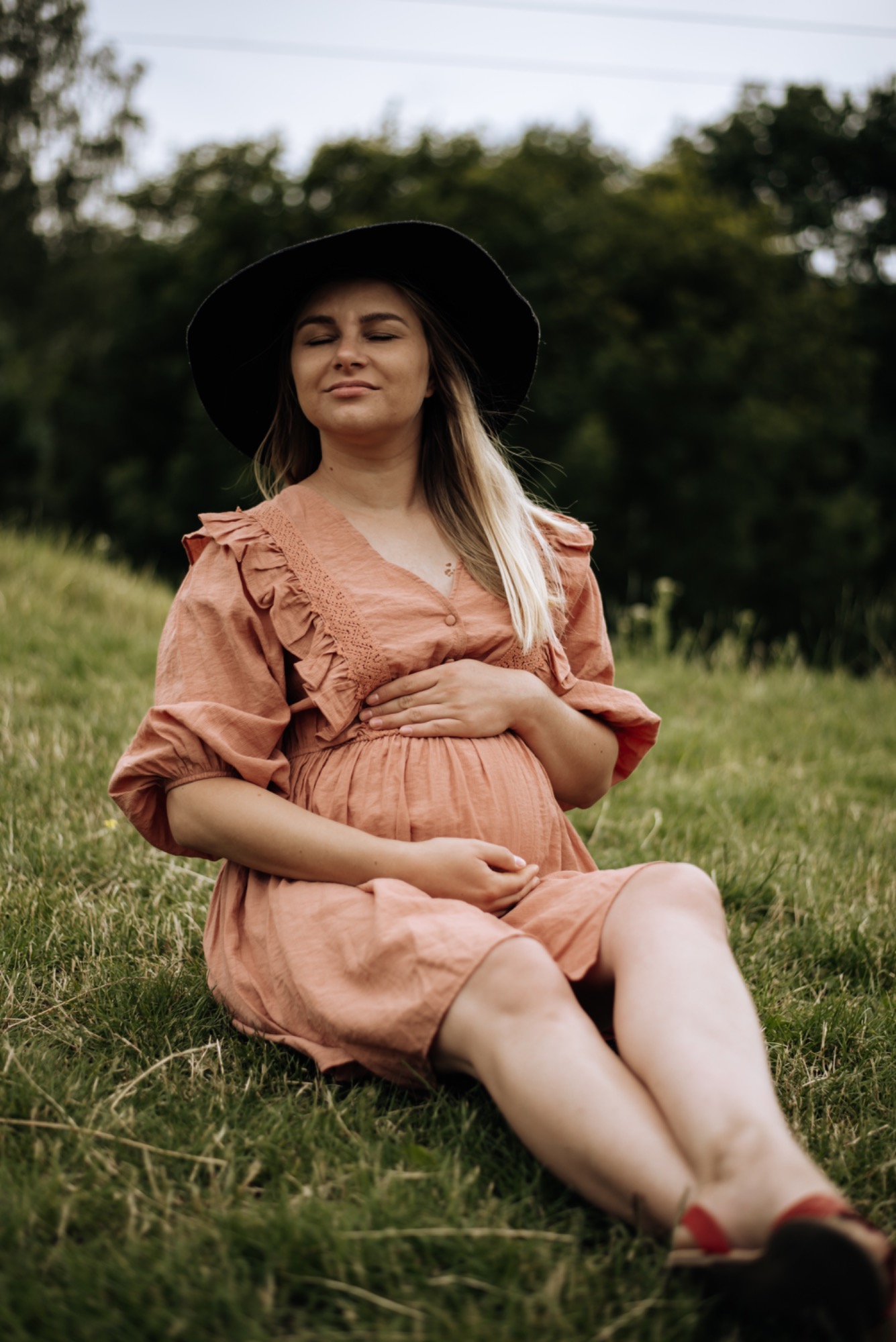 Kobieta w ciąży w zwiewnej sukience i kapeluszu siedzi na trawie z zamkniętymi oczami, dłońmi obejmując brzuch, w tle drzewa.