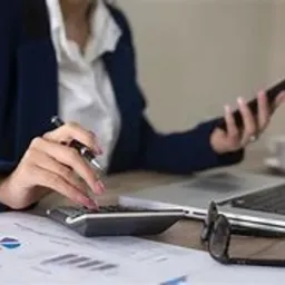 Kobieta w granatowym żakiecie oblicza dane na kalkulatorze, trzymając długopis w dłoni, z rozłożonymi wykresami i okularami na biurku.