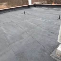 Płaski dach pokryty papą termozgrzewalną z widocznymi kominkami wentylacyjnymi i kominem w tle, zabezpieczony obróbką blacharską.