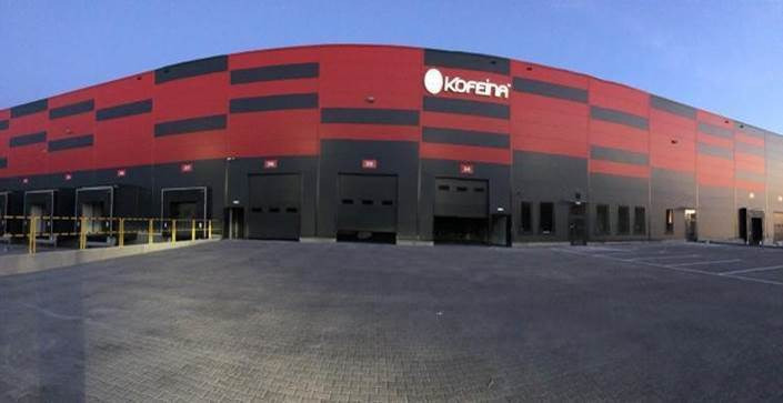 Szeroki kadr na nowoczesny magazyn w kolorach czerwieni i szarości, z widocznym logo 'KOFEINA' nad czarnymi bramami wjazdowymi i rozległym placem manewrowym.