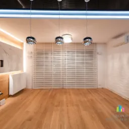 Nowoczesne wnętrze pokoju po wykończeniu: drewniana podłoga, ściana z panelami imitującymi drewno i marmur z podświetleniem, biała ściana z dekoracyjną fakturą, żaluzje poziome w oknie...