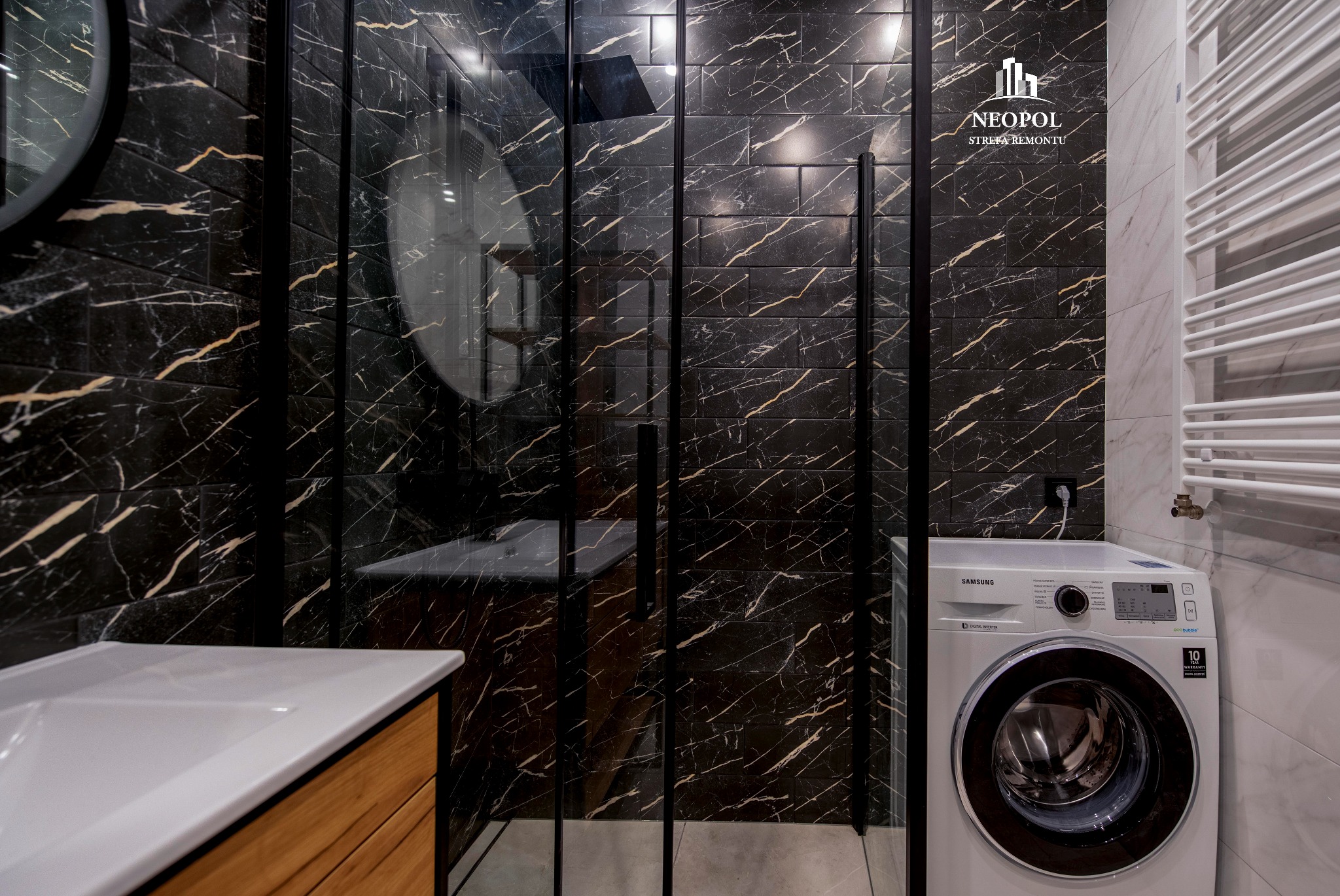 Nowoczesna łazienka z czarnymi płytkami imitującymi marmur, kabiną prysznicową typu walk-in z czarnymi profilami, umywalką na drewnianej szafce i pralką Samsung.