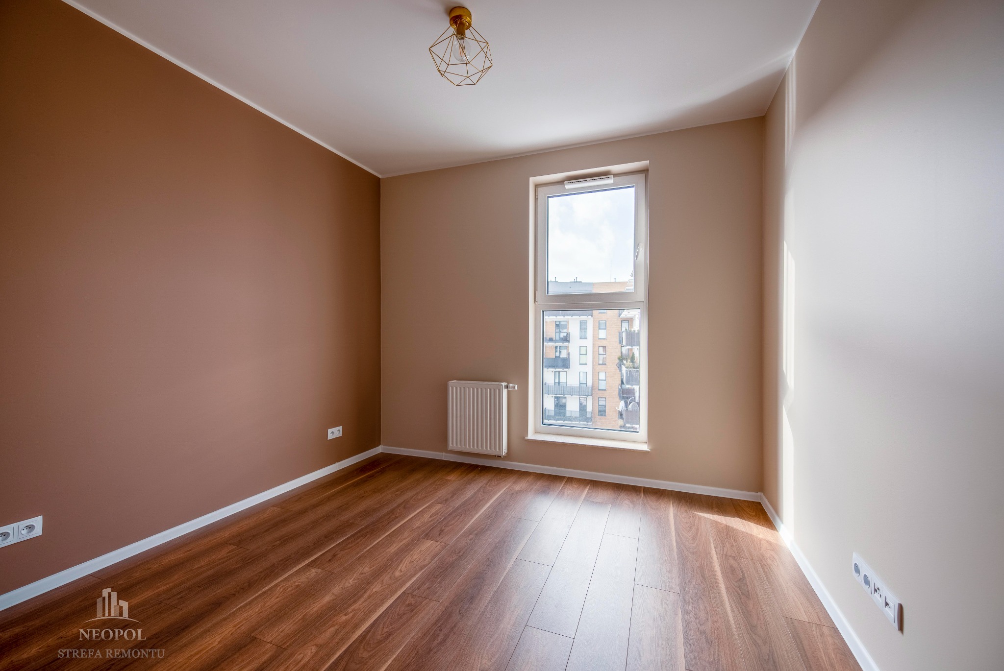 Pusty pokój po odświeżeniu, z brązową ścianą, drewnopodobną podłogą, białym oknem z widokiem na budynek i geometryczną lampą na suficie.